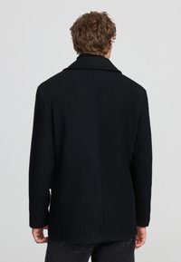 Uomo con capelli ricci che indossa un cappotto nero e jeans scuri, in piedi di spalle su uno sfondo grigio semplice.