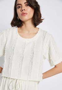 Weißer strukturierter, gekürzter Pullover mit ausgefransten Akzenten und runder Halslinie, kombiniert mit einem passenden Rock mit Kordelzug in der Taille.