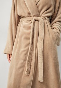 Beige, pluizige badjas met een schouderkraag, een ceintuur in de taille en zijzakken. De zachte structuur en lange mouwen zorgen voor comfortabele bedekking.