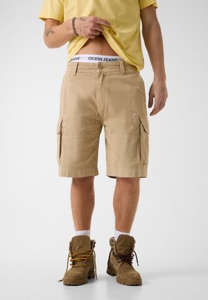 Mężczyzna podnoszący żółtą koszulkę, aby pokazać biały pas od spodni Guess Jeans, ubrany w beżowe szorty cargo i brązowe buty sznurowane z białymi skarpetami.