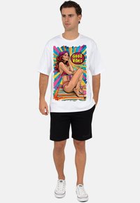 Wit grafisch T-shirt met kleurrijk "Good Vibes" ontwerp van een vrouw in een bikini, gecombineerd met zwarte shorts en witte sneakers.