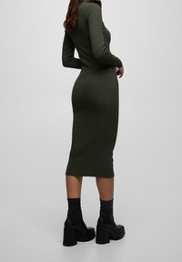 Femme portant une robe midi olive verte ajustée à manches longues et des bottines à plateforme noires sur un fond uni.