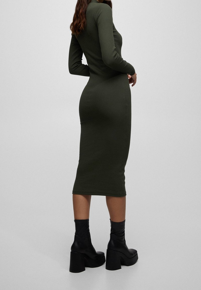 Femme portant une robe midi olive verte ajustée à manches longues et des bottines à plateforme noires sur un fond uni.