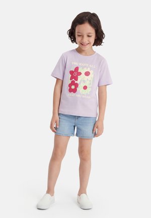 Šypsosi mergina trumpais tamsiais plaukais, vilkinti levandų spalvos marškinėlį su gėlėmis ir užrašu, džinsinius šortus ir baltus slip-on batus.