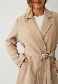 Beige wikkeljas met een strikceintuur, brede revers en twee voorzakken. Gemaakt van gestructureerde stof met een gladde afwerking.
