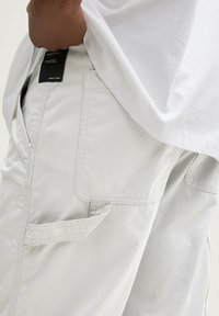 Pantaloni cargo grigi chiari con numerose tasche, cuciture visibili e un'etichetta sulla vita. Il tessuto presenta una leggera trama.