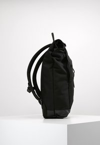 Mochila negra con diseño de parte superior enrollable, tela texturizada, correas acolchadas y base resistente. Cuenta con un bolsillo lateral y hardware mínimo.