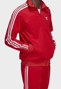 Red athletic tracksuit with white three-stripe accents. Zippered jacket features a stand-up collar and side pockets. Smooth fabric texture.