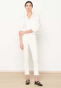 Camisa blanca de botones con mangas enrolladas, combinada con jeans blancos ajustados. Zapatos tipo slip-on negros con tachuelas plateadas. Diseño simple y limpio.
