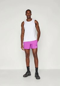 Biely tielko, ružové šortky s logom Nike, čierne športové topánky, stojí vzpriamene na neutrálnom pozadí. Hladká textúra a priliehavý dizajn.