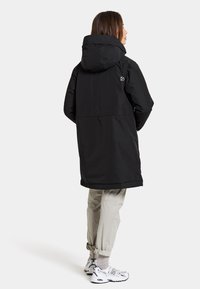 Parka nera isolata con cappuccio, caratterizzata da un design snello, tasche laterali e un dettaglio del logo sulla manica. Abbinata a pantaloni leggeri.