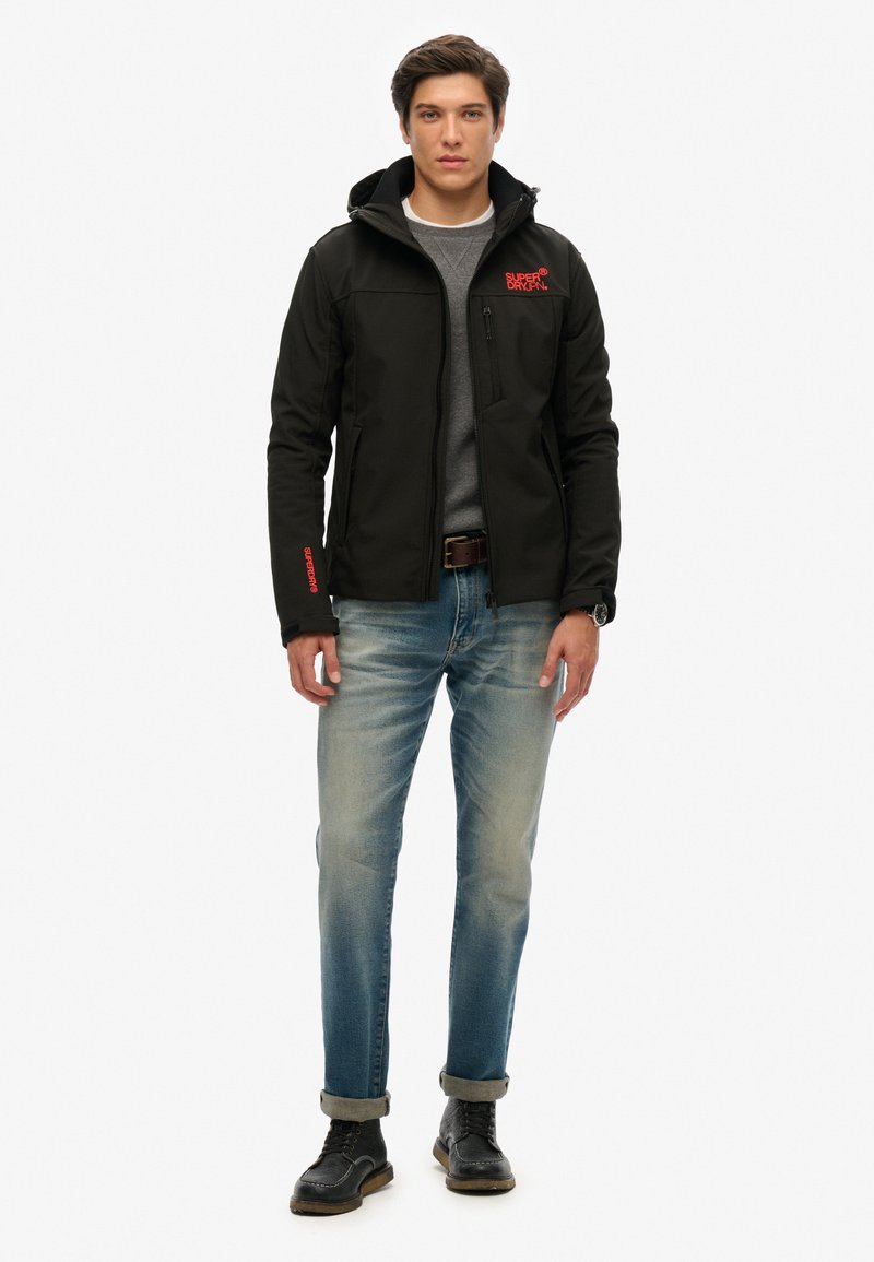 Zwarte softshell jas met een capuchon, ritssluitingen aan de voorkant en een rood logo op de borst. Gecombineerd met lichtblauwe jeans en zwarte schoenen.