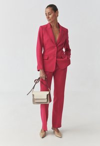 Přiléhavý jasně růžový blazer a odpovídající kalhoty z hladkého materiálu. Béžová kabelka s klopou a popruhem. Modelka v tělových lodičkách.