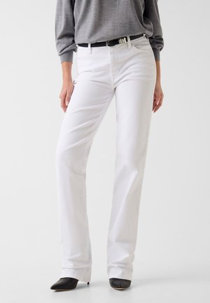 Kobieta ubrana w białe proste spodnie z wysokim stanem, czarny pasek, szara bluzka z długim rękawem oraz czarne czółenka z ostrym noskiem, stojąca na białym tle.