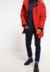 Parka isolée rouge avec une capuche doublée de fausse fourrure, fermetures éclair et boutons ; associée à un pull à motifs bleu foncé et un jean noir. Bottines noires.