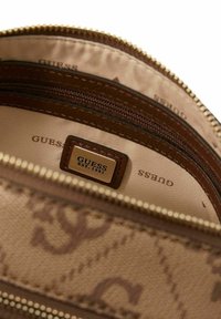 Portefeuille marron à motifs avec ferrures dorées et poches intérieures zippées. L'intérieur présente une doublure en tissu avec le logo "GUESS" répété.