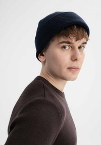 Marineblauwe geribbelde gebreide beanie gedragen door een model met lichtbruin haar, gecombineerd met een donkerbruine crewneck trui, tegen een lichte achtergrond.