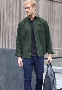 Camisa de cuadros verdes, con una camisa negra debajo, combinada con jeans azul oscuro. El modelo sostiene un bolso de cuero negro, apoyado contra un fondo texturizado.