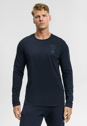 Junger Mann mit kurzen blonden Haaren, der ein eng anliegendes marineblaues Langarmshirt mit dezentem Text auf Brust und Ärmeln trägt und vor einem schlichten Hintergrund steht.