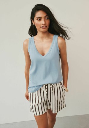 Frau mit langen dunklen Haaren, die ein hellblaues ärmelloses Oberteil und weiße Shorts mit vertikalen schwarzen Streifen in einer neutralen Umgebung trägt.