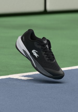 Chaussure de sport noire avec semelle blanche et logo flottant au-dessus d'une surface de court de tennis bleue et verte.