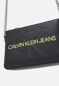 Černá crossbody taška z umělé kůže s embosovaným logem. Odvážný neonově žlutý text zní "CALVIN KLEIN JEANS." Vybavena stříbrným řetízkem.