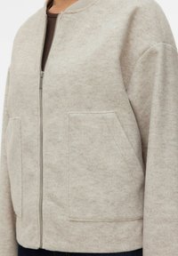 Veste en laine beige zippée avec col côtelé et deux grandes poches plaquées sur le devant, portée sur un haut marron foncé.