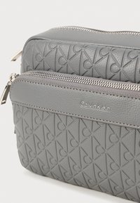 Grå tekstureret crossbody taske med et quiltet mønster og sølvhardware, med et synligt logo på en glat accentstribe.