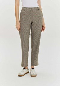 Kobieta w wysokich, wąskich spodniach w kratę i białych sneakersach, stojąca na tle jednolitego białego tła.