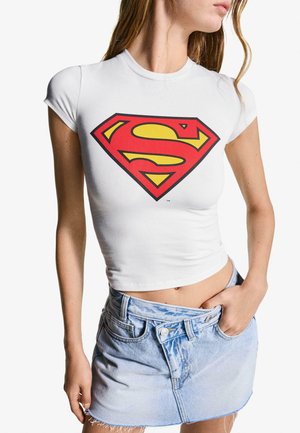 Hvid, tætsiddende T-shirt med korte ærmer, der har et stort rødt og gult Superman-logo på forsiden, parret med en lyseblå denim miniskørt.