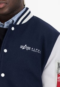 Námornícky modrá vlnená bomber bunda s bielymi rukávmi, zapečaťovacími gombíkmi, vyšívaným logom "Alpha Industries" a detailom na zips vrecka.