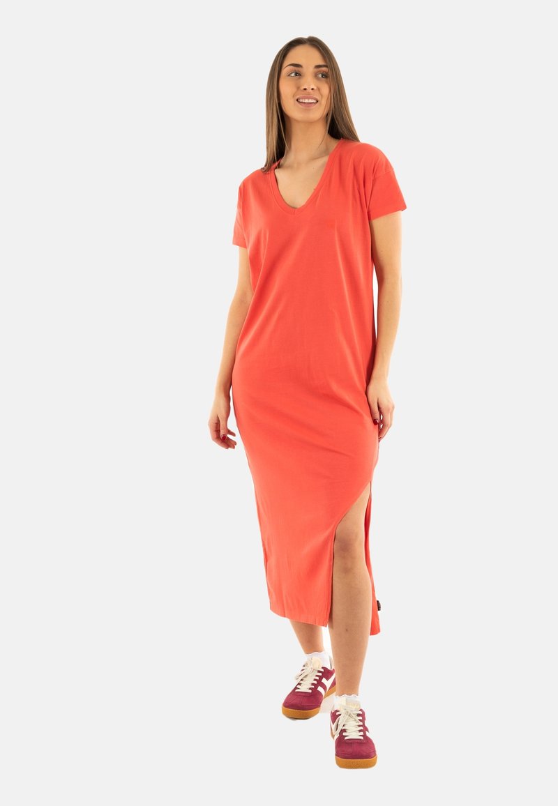 Robe longue corail à manches courtes, décolleté en V profond, fentes latérales, confectionnée dans un tissu doux, associée à des baskets bordeaux et blanches.