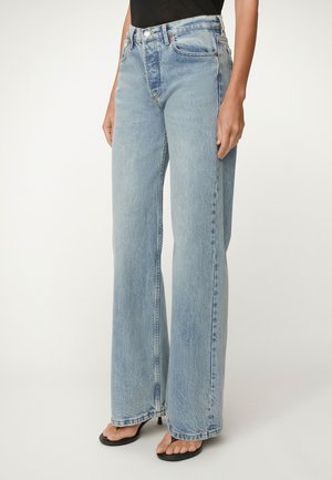 Model draagt lichtblauwe jeans met hoge taille en wijde pijpen, gecombineerd met zwarte open teen sandalen met hak, staand tegen een effen achtergrond.