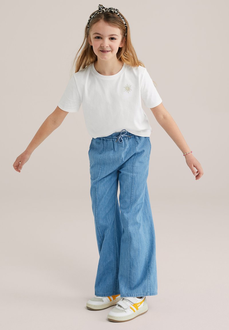 Camiseta blanca de algodón con un gráfico del sol; pantalones de mezclilla anchos azules; zapatillas blancas con detalles amarillos; el modelo lleva una diadema estampada.
