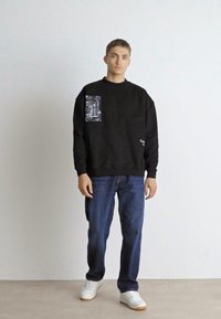 Zwarte sweatshirt met een grafisch zakontwerp, ronde hals, geribde zoom en manchetten, gecombineerd met blauwe jeans en witte sneakers.