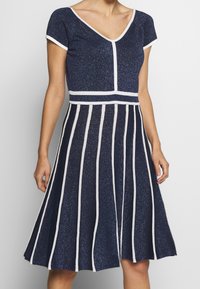 Abito in maglia glitter blu navale con scollatura a V, maniche corte e righe orizzontali bianche; silhouette svasata, lunghezza ginocchio, tessuto testurizzato.