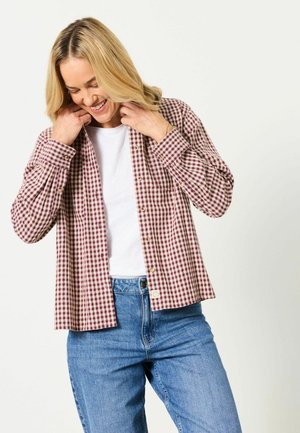 Lyshåret kvinde iført blå jeans, hvid t-shirt og en åben rød- og hvidternet skjorte, smilende og justerende sin krave.