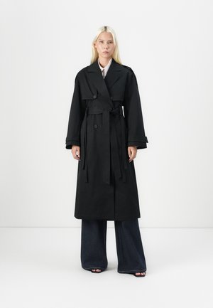 Woman with long blonde hair wearing a black belted trench coat, wide-leg dark jeans, and black open-toe shoes against a white background.