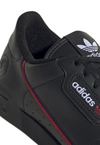 Melnas ādas sporta apavi ar sarkanu šuvju, mīkstu apkakli, teksturētu virsmu un ikonisko baltu adidas logotipu ar trim svītrām.