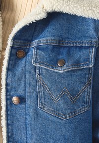 Jeansowa kurtka z białym sztucznym futrem przy kołnierzu, z miedzianymi guzikami i charakterystycznym haftem „W” na kieszeni na piersi. Widać niebieską teksturę dżinsu.