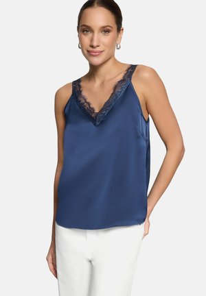 Kobieta ubrana w granatowy satynowy top bez rękawów z koronkowym dekoltem w kształcie litery V i białe spodnie, pozująca na tle jasno szarym.