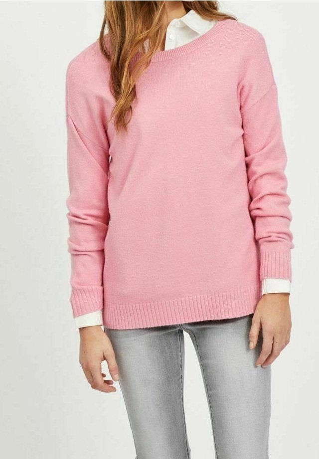 Femme portant un pull en maille rose clair par-dessus une chemise blanche à col, associée à un jean gris clair, mains détendues le long du corps.