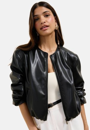 Naine kannab kuldseid kõrvarõngaid ja musta tehisnahanööpkattega jopet valge topi ja valgete pükstega musta vööga.