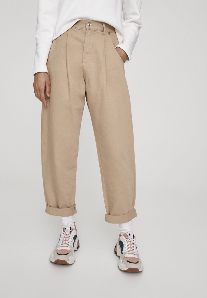Pantalones beige de cintura alta con piernas anchas, pliegues frontales, puños enrollados y bolsillos laterales. Combinados con zapatillas deportivas coloridas.