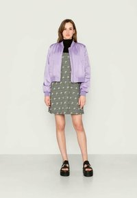 Casaco bomber lilás feito de material brilhante, usado sobre um vestido de padrão xadrez. Sandálias plataforma pretas completam o look.