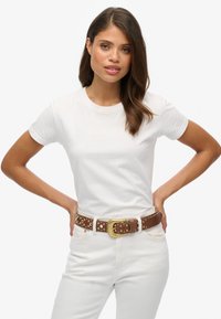 Weißes kurzärmliges T-Shirt kombiniert mit einem braunen, mit Nieten besetzten Gürtel mit goldener Schnalle; das Model posiert mit den Händen auf den Hüften und trägt weiße Hosen.