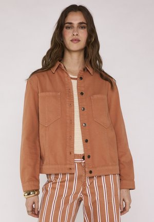 Donna che indossa una giacca marrone con bottoni sopra una maglietta beige e pantaloni a righe verticali marroni e bianchi, in piedi su uno sfondo chiaro.