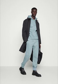 Lichtblauwe hoodie en sweatpants, gedragen onder een donkergrijze jas. Zwarte rubberen laarzen met een gestructureerde zool en een zwarte tote bag.