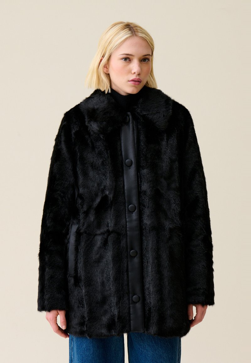 Cappotto in pelliccia sintetica nera con chiusura a bottone lucido nero, colletto oversize e tasche laterali. Presenta un aspetto morbido e testurizzato.