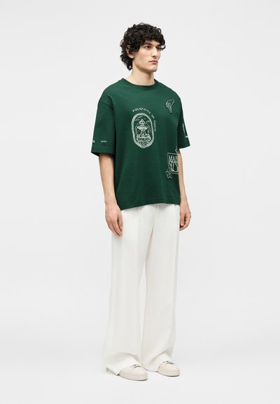 Νέος άνδρας που φοράει σκούρο πράσινο t-shirt με γραφικά, λευκό παντελόνι με φαρδιά μπατζάκια και μπεζ αθλητικά παπούτσια, στέκεται μπροστά σε λευκό απλό φόντο.